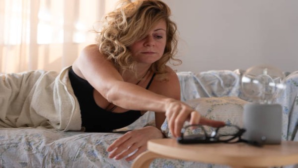 Seorang wanita mematikan alarm pagi sambil terlihat mengantuk, ilustrasi tantangan bangun terlalu dini bagi tubuh yang belum siap.