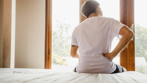 Seorang pria tampak memegang punggung bagian bawah saat duduk di tepi tempat tidur akibat nyeri otot dan kelelahan berkepanjangan.